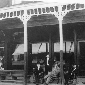 Group of white men in suits smoking in rocking chairs and boy on bench on covered hotel porch