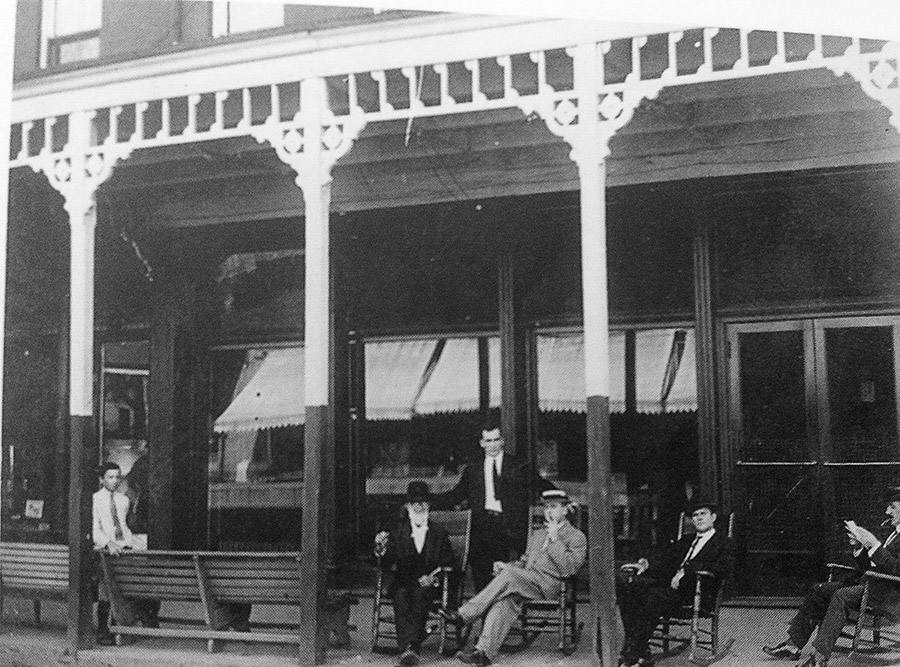 Group of white men in suits smoking in rocking chairs and boy on bench on covered hotel porch