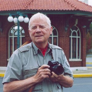Older white man with camera standing outside brick building with arched windows and street lamps