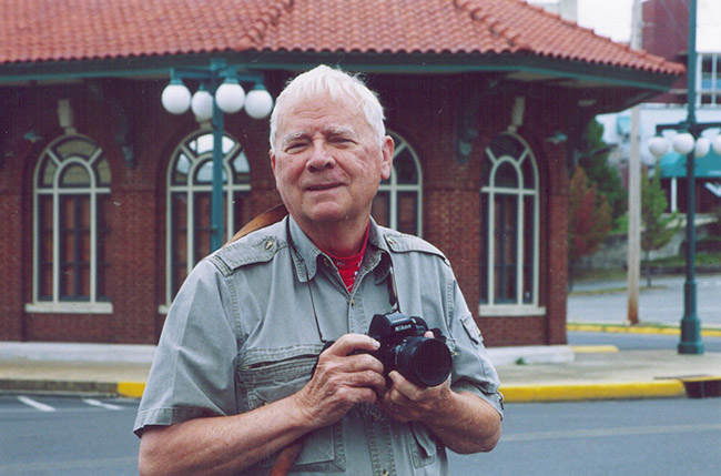 Older white man with camera standing outside brick building with arched windows and street lamps