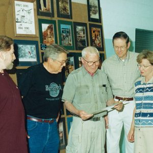 White men and woman standing together looking at photograph with framed pictures on the wall behind them