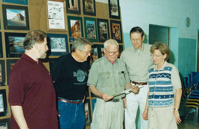 White men and woman standing together looking at photograph with framed pictures on the wall behind them