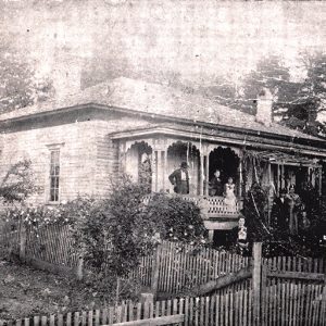 White man woman and children outside single-story house with covered porch and fence