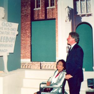 White man and African-American woman in wheelchair looking at statue of a young woman holding a sign that reads "God gave his only son for the freedom of mankind NAACP"