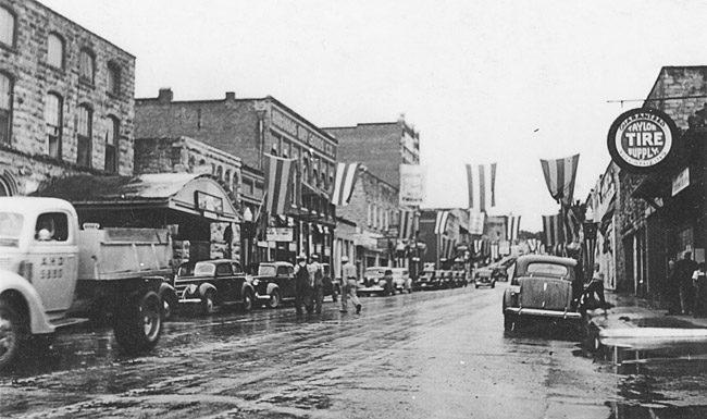 rainy street lined with buildings and parked cars with striped flags hanging above it