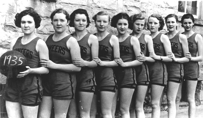 Group of young white women in sleeveless shirts and shorts standing in line against a brick wall with ball