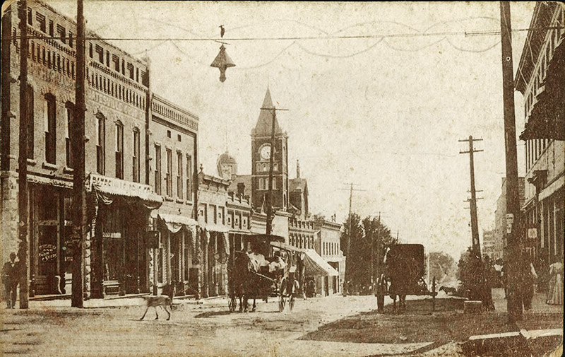 Horse drawn carriages on street between rows of multistory brick buildings with clock tower in the background