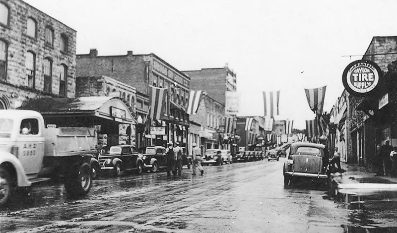 Parked cars on wet street with buildings on both sides and flags hanging above it