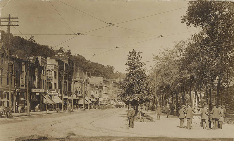 Street with row of multistory buildings and power lines on its left side and crowd of men in suits on its right side