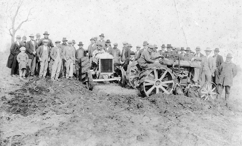 Crowd of men posing with two tractors in a field