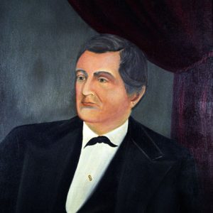 White man in black suit with red curtains in the background
