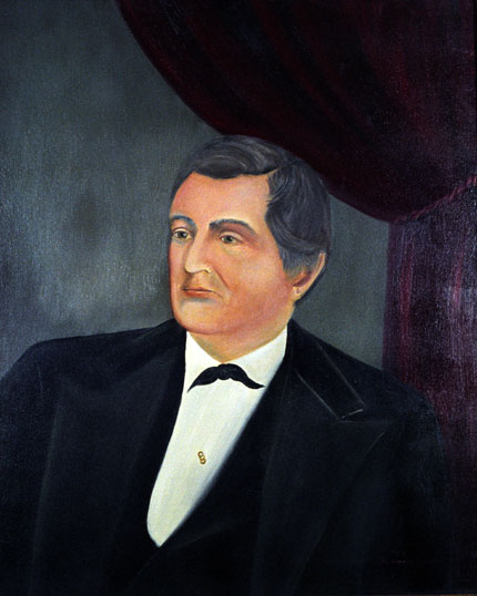 White man in black suit with red curtains in the background