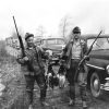 two men dressed for the outdoors holding shotguns and dead ducks in front of a row of old cars parked in a field