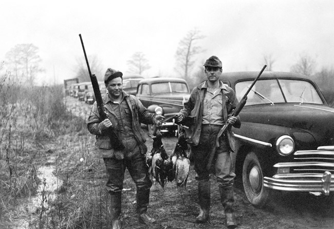 two men dressed for the outdoors holding shotguns and dead ducks in front of a row of old cars parked in a field