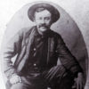 White man with a mustache and hat sitting on a barrel