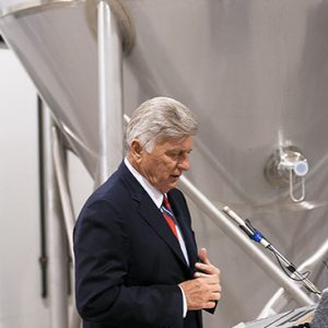 White man with gray hair in suit speaking into a microphone at lectern next to brewing machinery