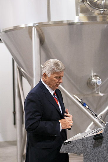 White man with gray hair in suit speaking into a microphone at lectern next to brewing machinery