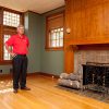 older white man in red shirt and dark pants standing in well lit room with wood floor fireplace and large windows