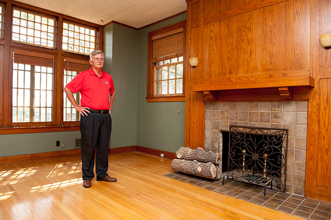 older white man in red shirt and dark pants standing in well lit room with wood floor fireplace and large windows