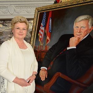 white woman dressed in white poses next to painting of white man in suit and tie posing in leather-backed chair with Arkansas and U.S. flags