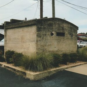 Small concrete building on parking lot with car parked in front of building in the background