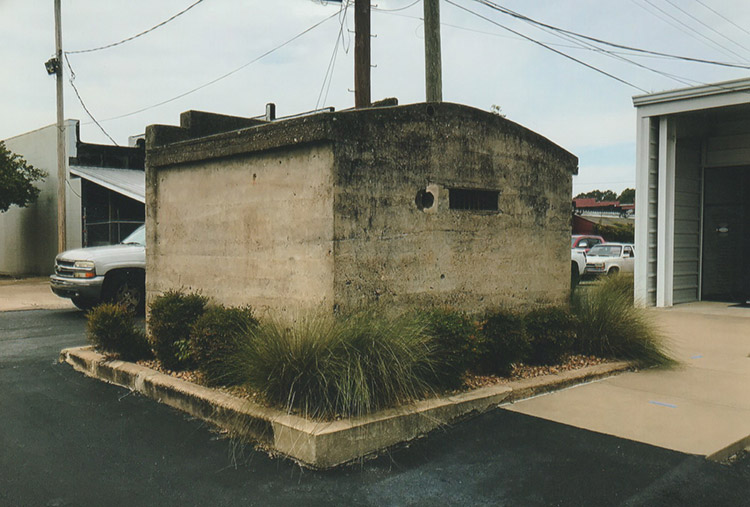 Small concrete building on parking lot with car parked in front of building in the background