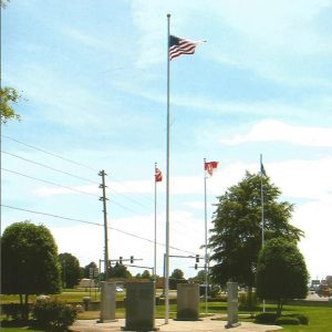 Engraved stone monuments arranged in circle formation with flag poles in the center with trees and traffic lights in the background