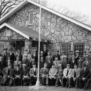 Group of white men in suits hats and topcoats outside building with stone walls and flag pole