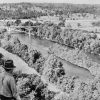 White man wearing a fedora hat looking over a tree-lined river and bridge in the distance