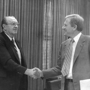 Older white man with glasses in suit and tie shaking hands with white man in suit and tie