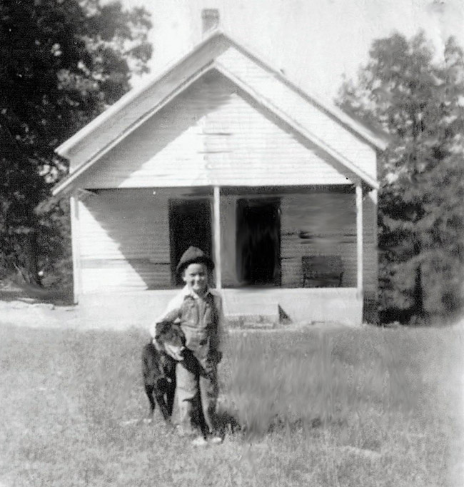 Young white child in overalls with dog standing in front of single-story building with two front doors