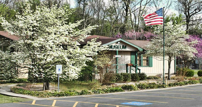 Single-story building with covered porch blooming trees and flag pole in front yard taken from parking lot