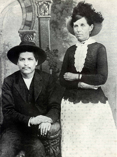 Cherokee man in hat and suit sitting next to white woman standing in hat and dress with her arms crossed