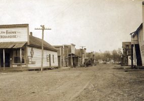 Dirt road with buildings on either side, including one "Dowell and Brown General Merchandise," and power lines