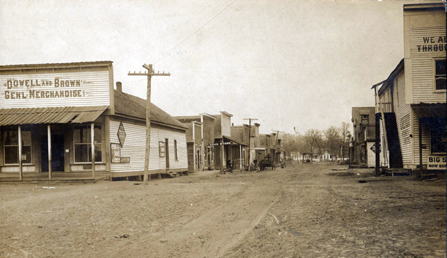 Dirt road with buildings on either side, including one "Dowell and Brown General Merchandise," and power lines