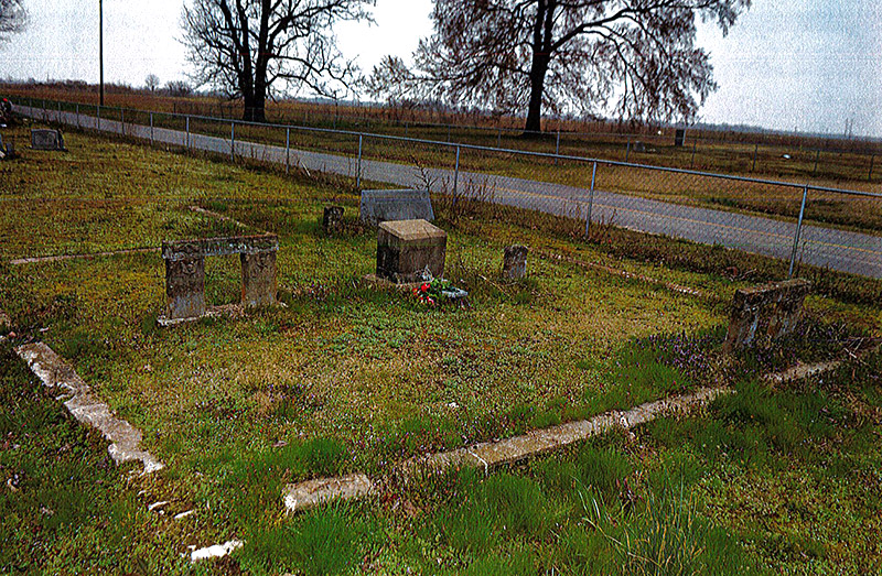 Gravestones in rural cemetery with fence next to road running alongside it
