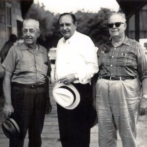 Three older white men in casual dress, on plank walkway near wood frame building and road