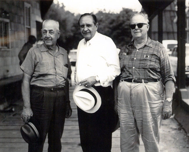Three older white men in casual dress, on plank walkway near wood frame building and road