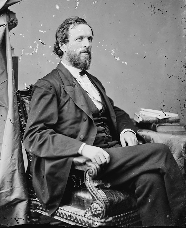 White man with beard in suit sitting in chair with stack of books on table next to him