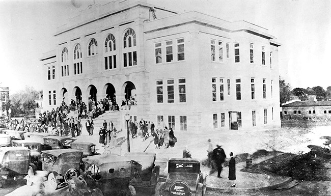 Crowd of people on steps of three-story building with parked cars