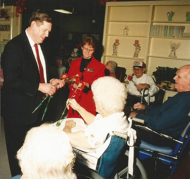 White man in suit handing roses to older white men and women