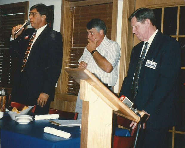 White man in suit with microphone speaking with white men standing next to him behind a long table