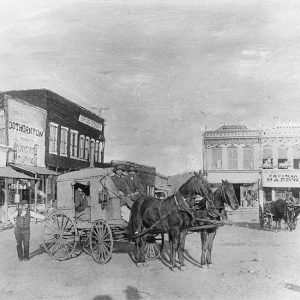Two two-horse stagecoaches in square with white drivers and multistory storefronts behind them
