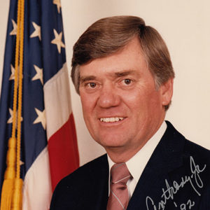 Autographed photograph of white man smiling in suit and tie with flag behind him