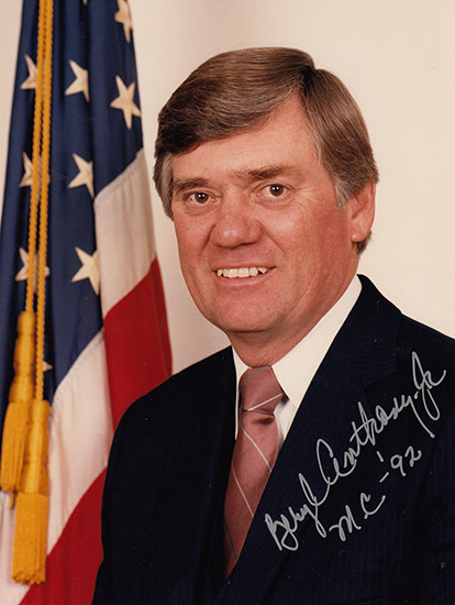 Autographed photograph of white man smiling in suit and tie with flag behind him