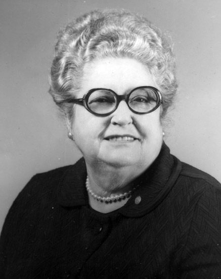 Older white woman with wavy pulled up hair and round glasses wearing a dark top and pearls