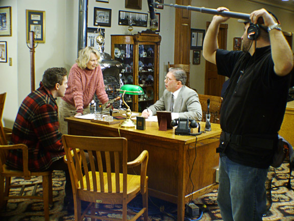White woman talking to young white man and older white man in office on film set with white boom microphone operator in the foreground