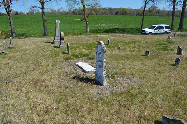 Stone monuments and gravestones in rural cemetery with green field next to it and car on driveway