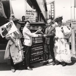 White men in suit buying jar of pickles from white man in suit and two white men in costumes outside Lowery store