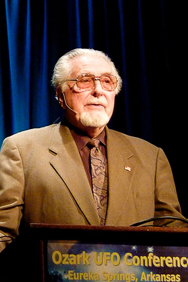 white man with white hair and goatee and tinted glass in brown suit speaking at lectern labeled "Ozark UFO Conference, Eureka Springs, Arkansas" in front of blue curtain